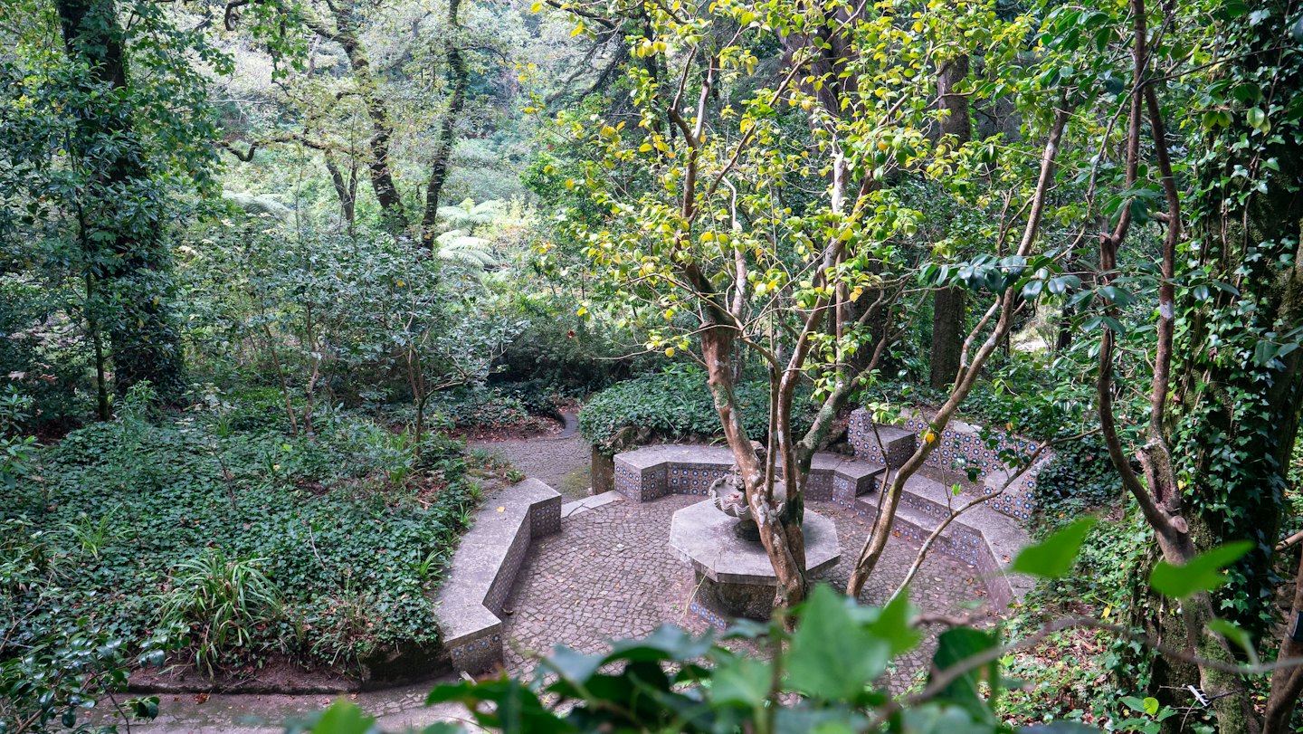 Secluded garden seating area with stone benches and a central tree.